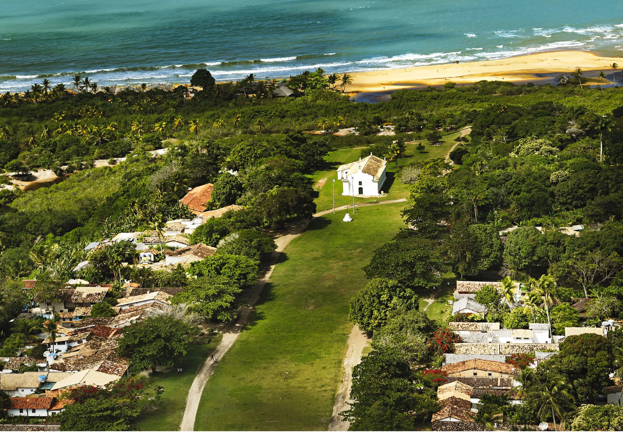 Aerial view of a coastal village with a white chapel on a green lawn among trees, beach and sea beyond