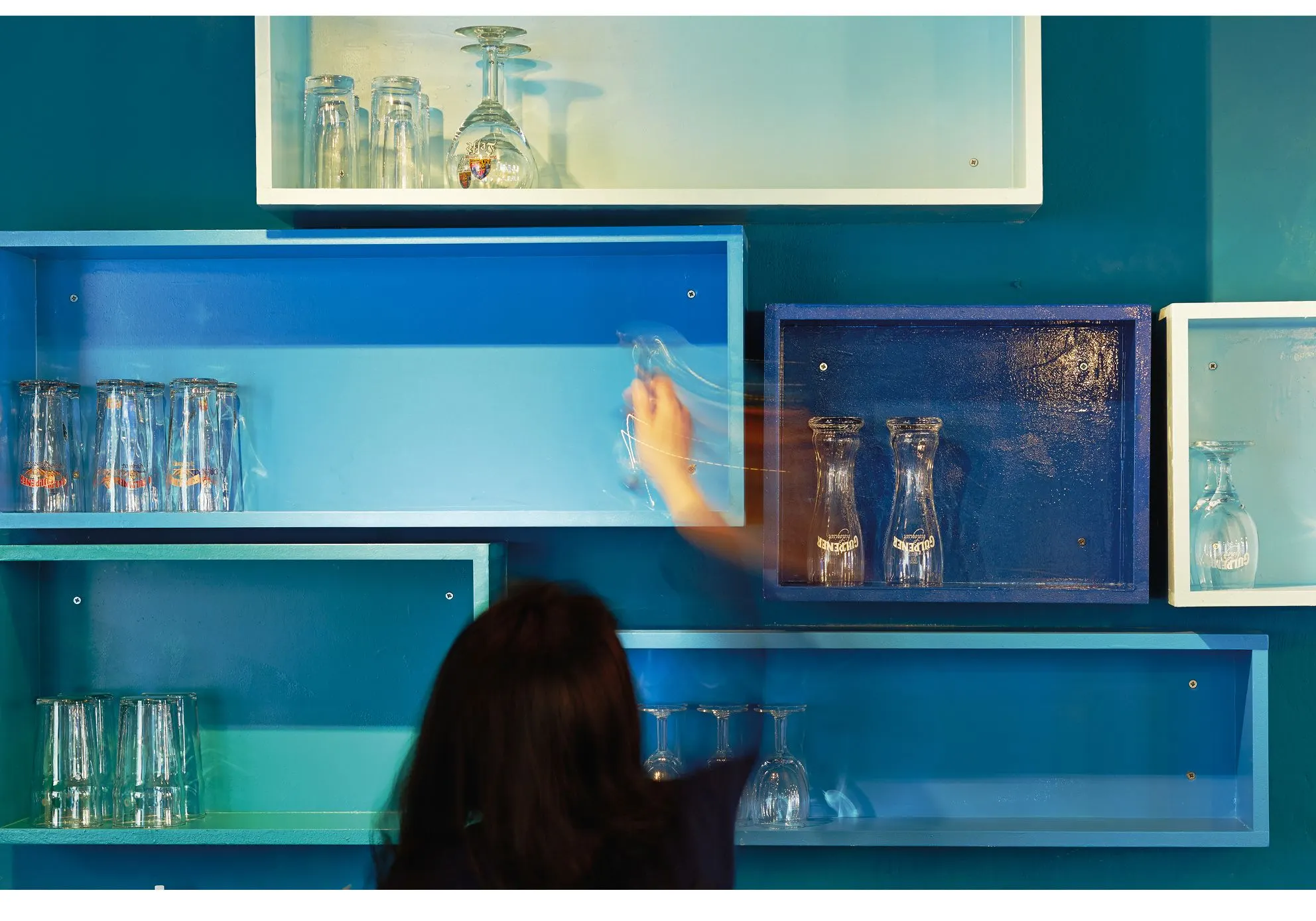 Person reaching for glasses on blue and turquoise wall shelves in the theatre bar