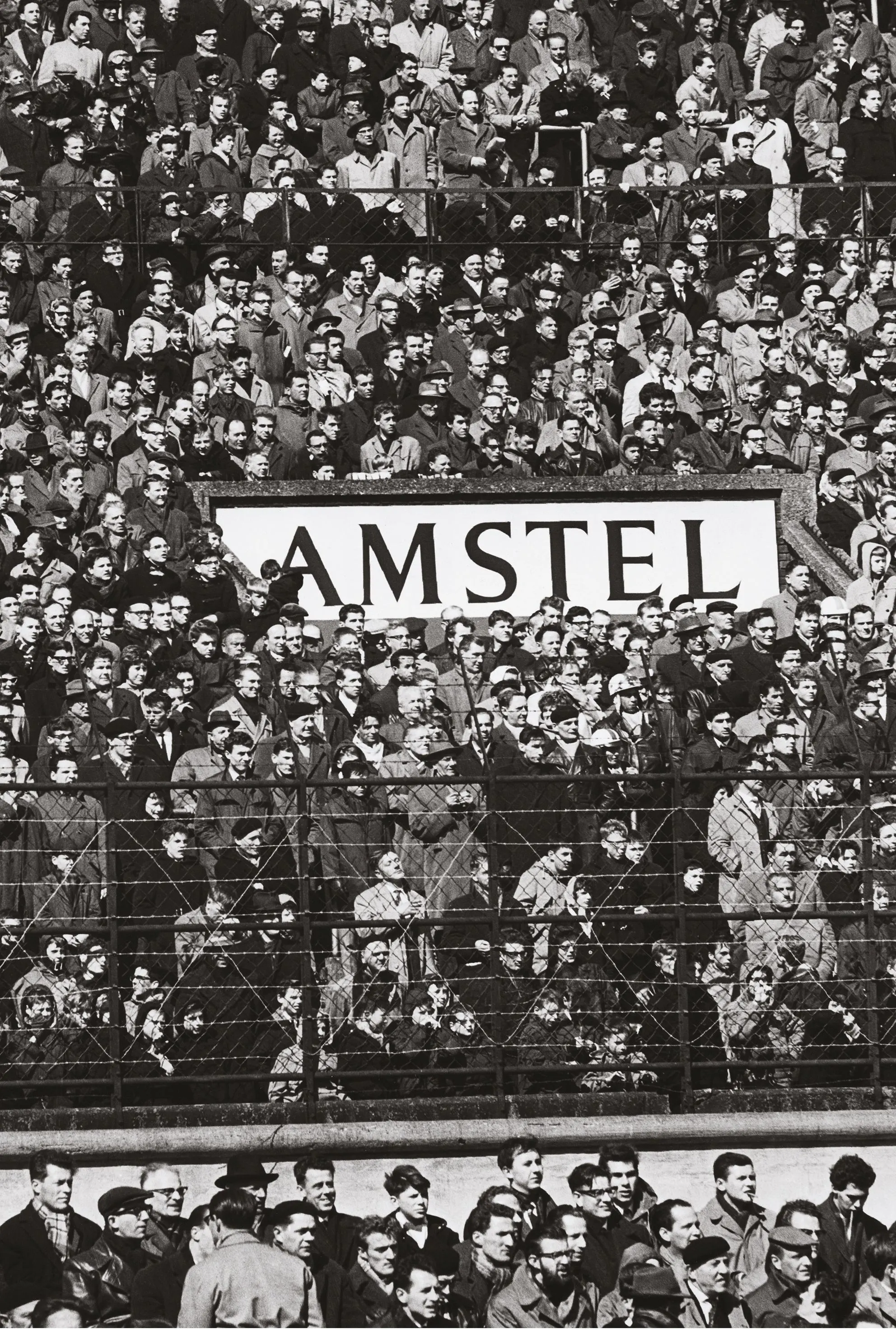 Black-and-white photo of a football stadium crowd behind a fence with an Amstel sign