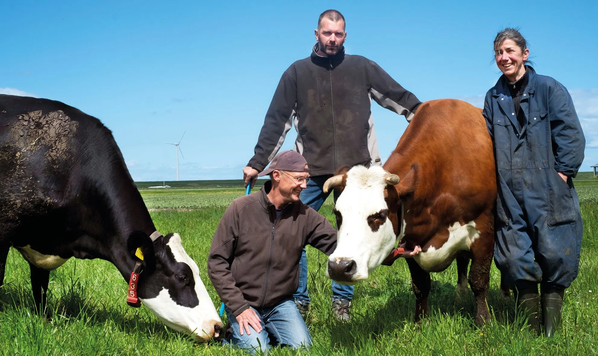 Two farmers and a woman posing with a black-and-white and a brown-and-white cow in a green pasture