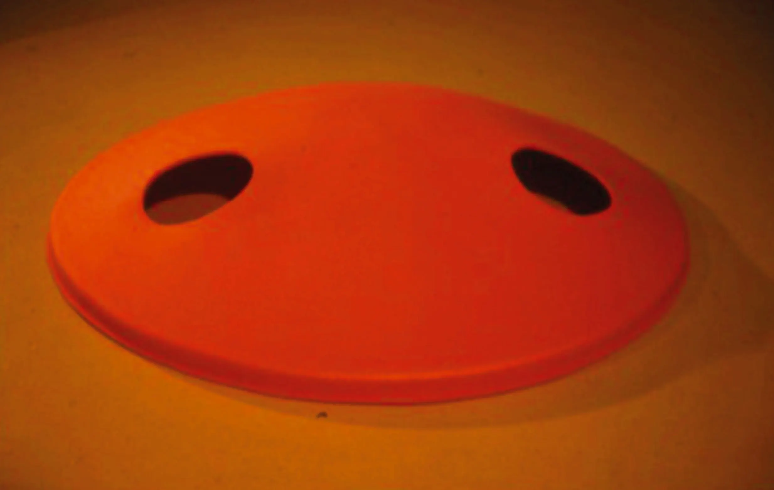 Red-orange disc-shaped soft sculpture with two oval holes on an orange background