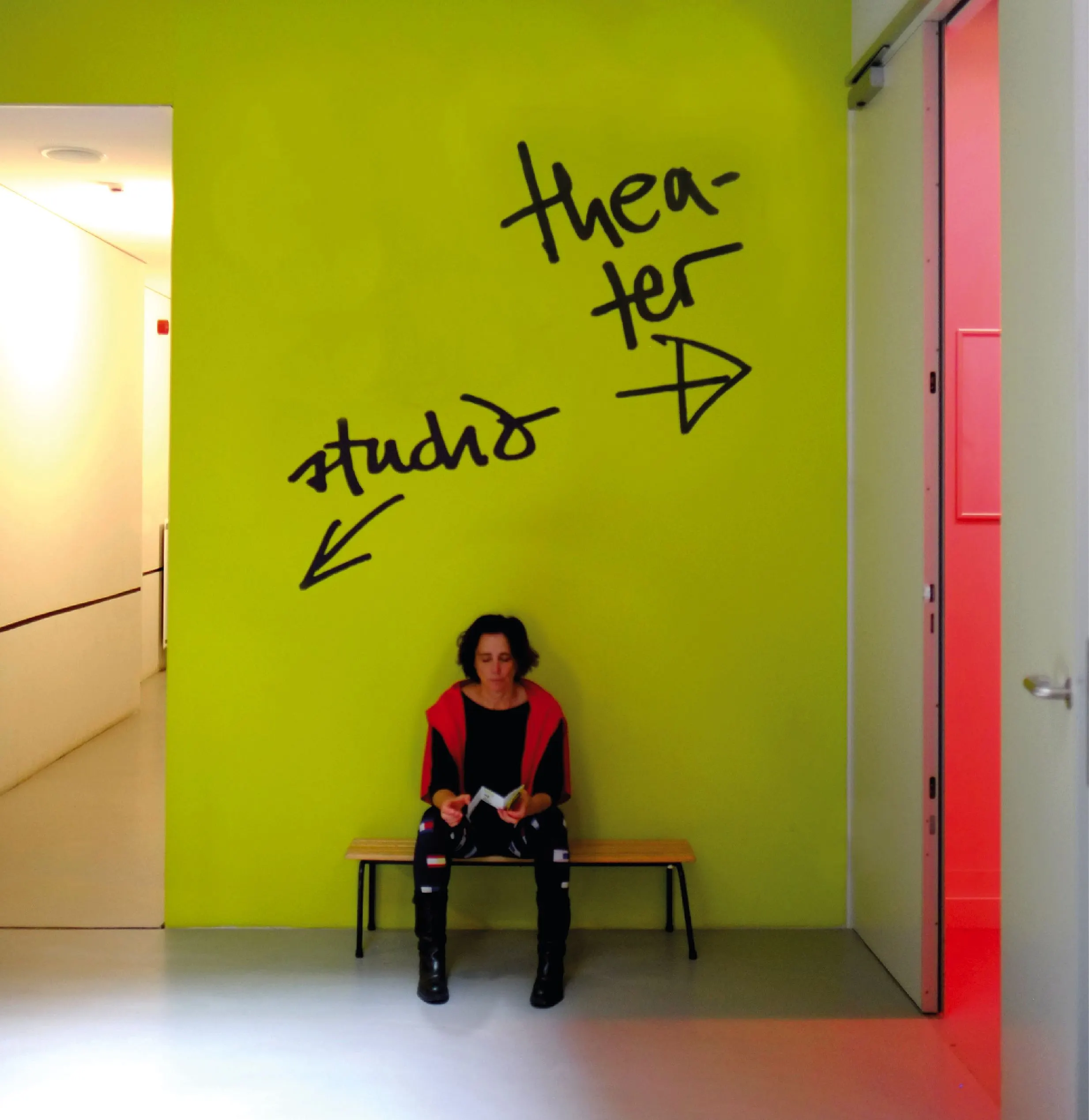 Bright yellow-green theatre corridor with handwritten wayfinding ‘theater’ and ‘studio’ arrows and a seated person