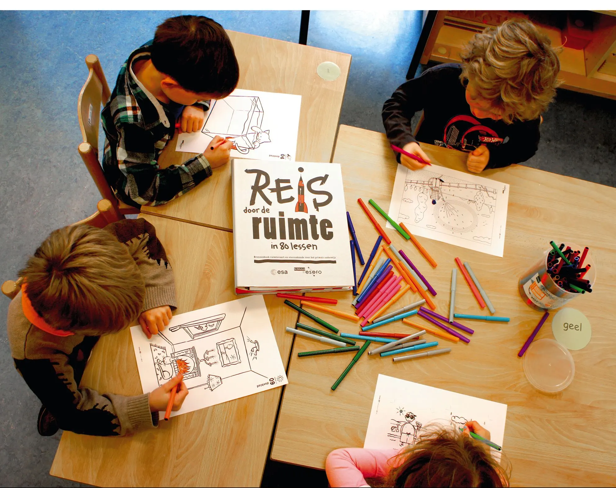 Overhead view of four children drawing with coloured pencils at tables around the ‘Reis door de ruimte’ binder