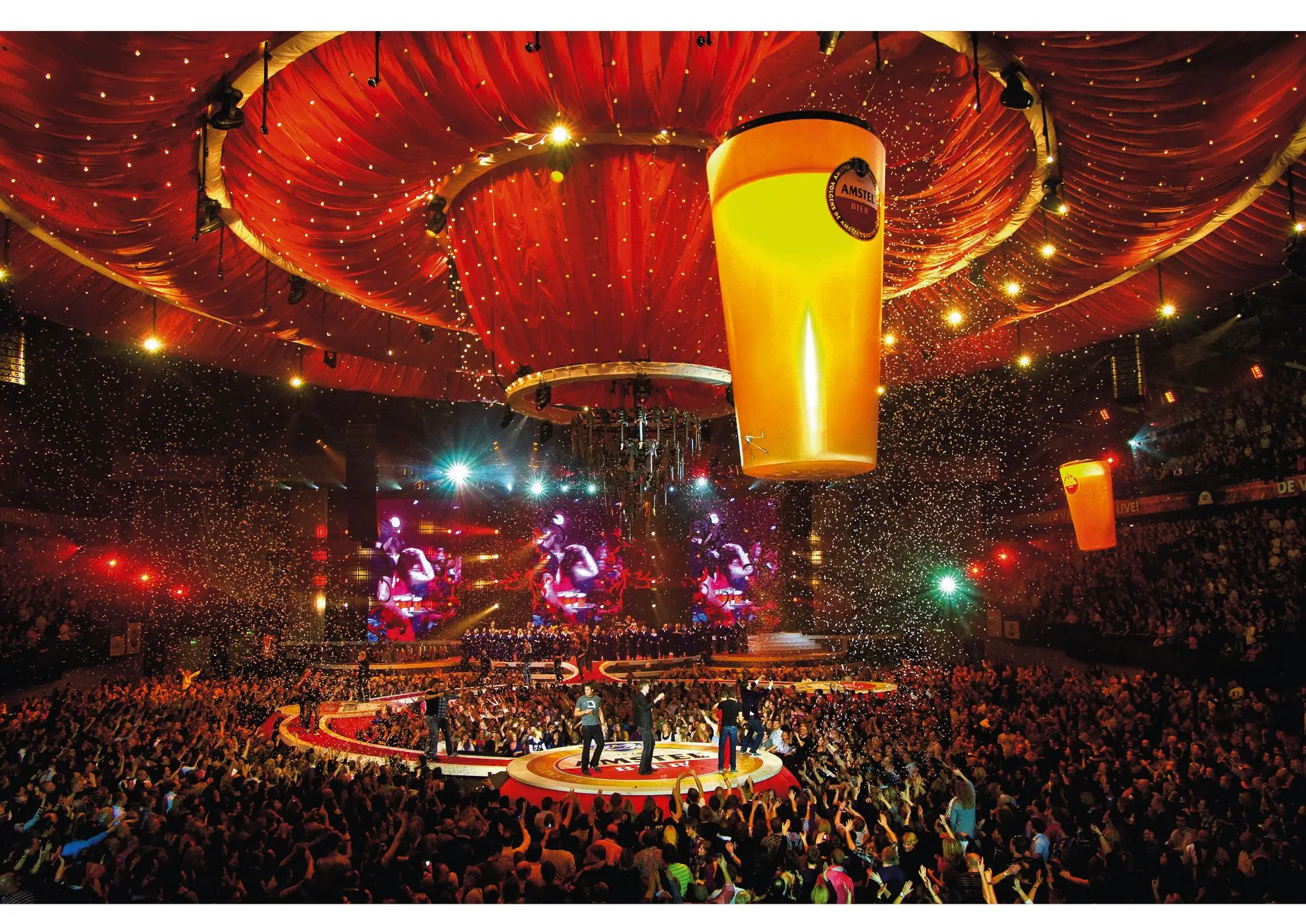 Packed concert arena in red light with a giant glowing beer glass hanging above the stage and confetti
