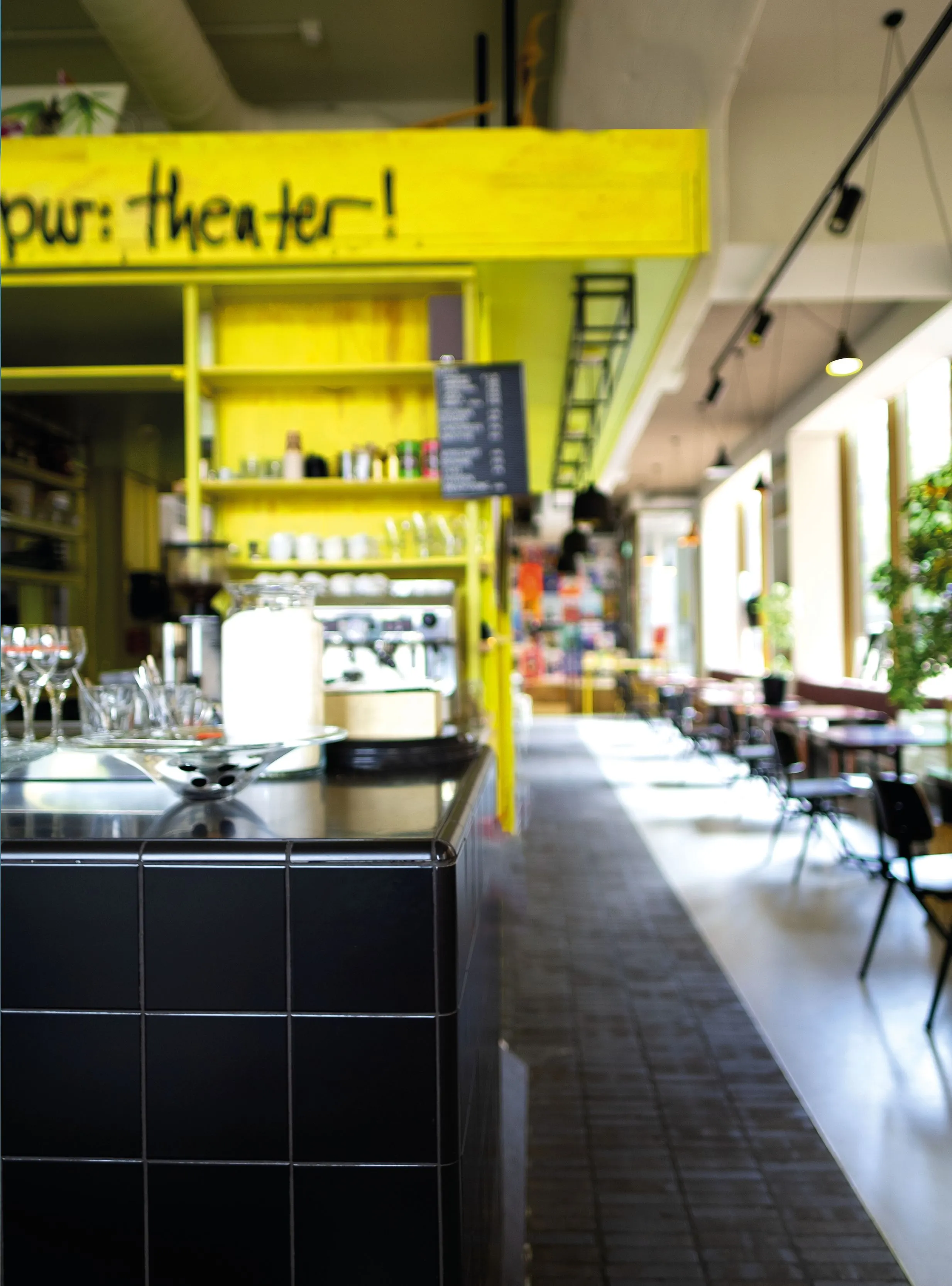 Theatre café with yellow shelving, black tiled counter and a long row of tables, sign reading ‘theater!’
