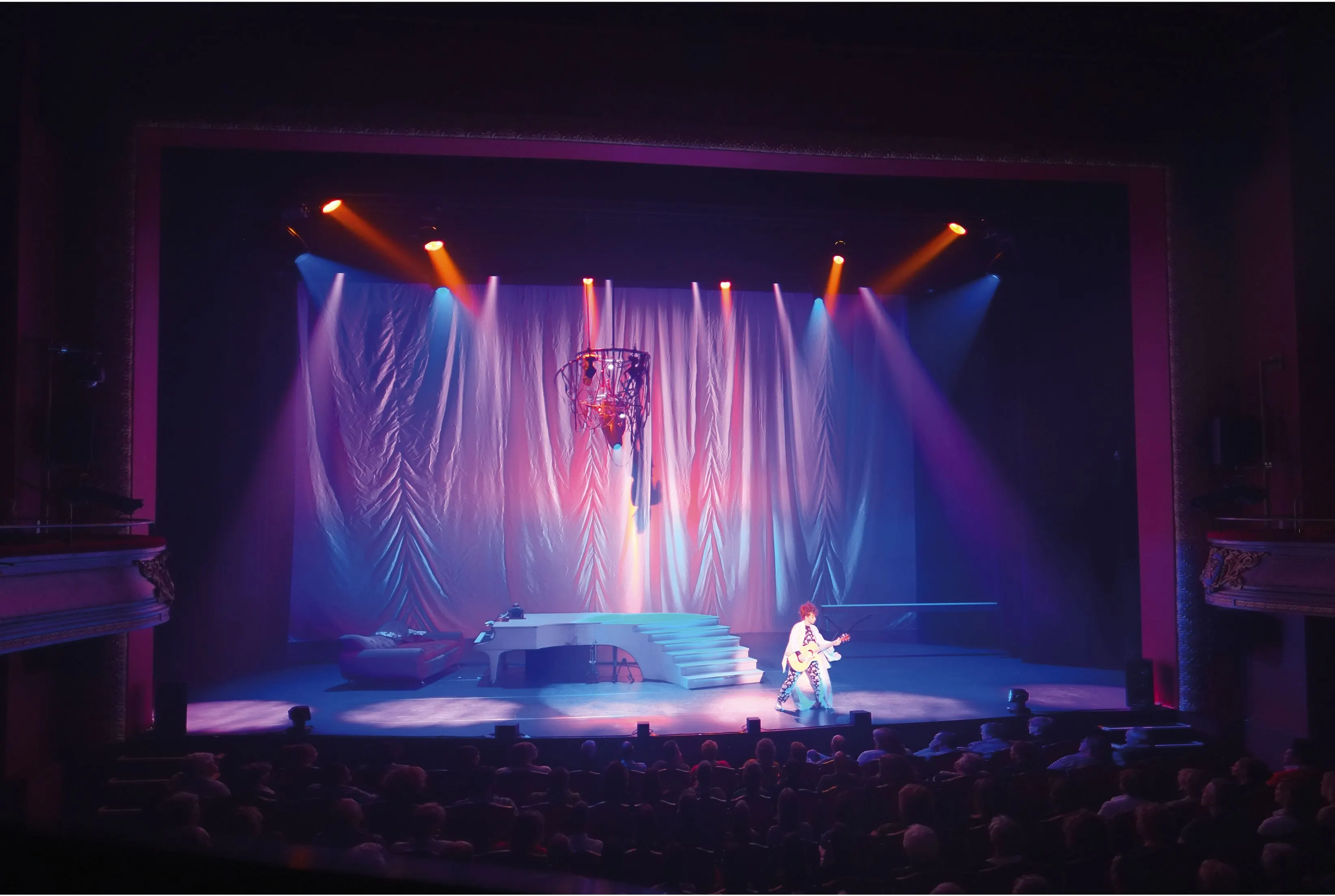 Theatre stage in blue and pink light with draped curtains, a grand piano on steps and a guitarist