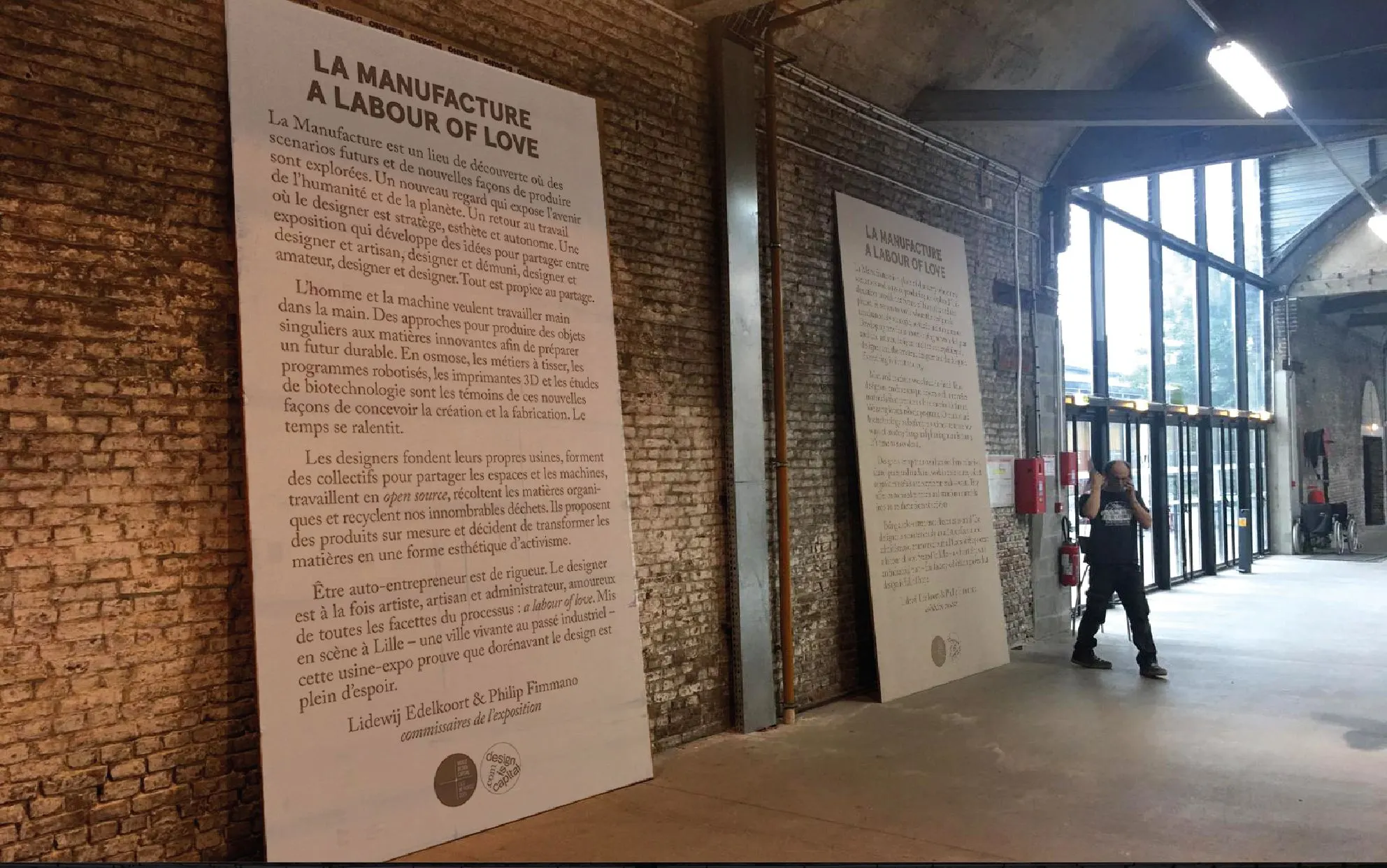 Large exhibition text panels for 'La Manufacture, A Labour of Love' in a brick industrial hall with tall windows