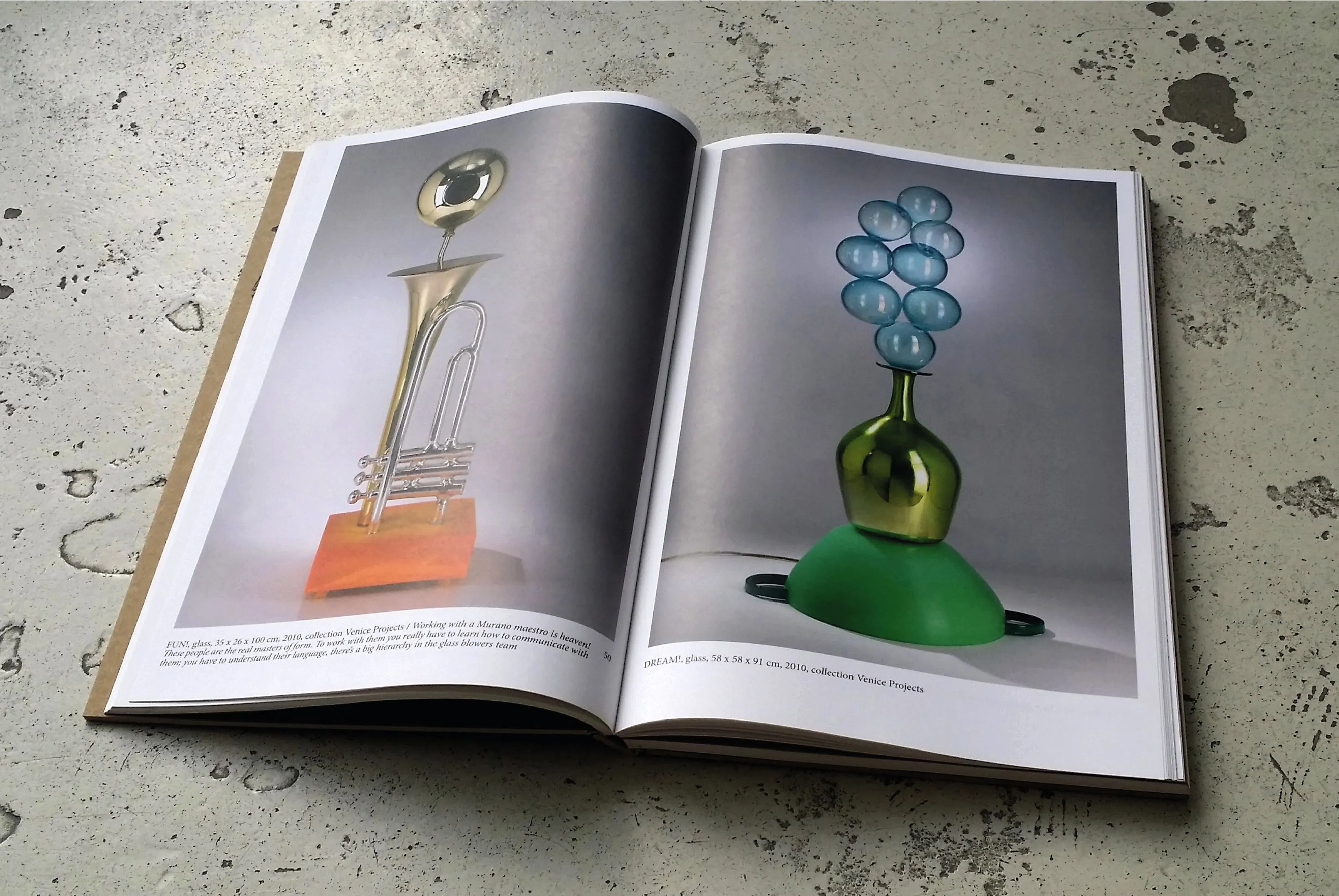 Open book on a concrete surface showing a brass trumpet-like object and a green and blue glass vase