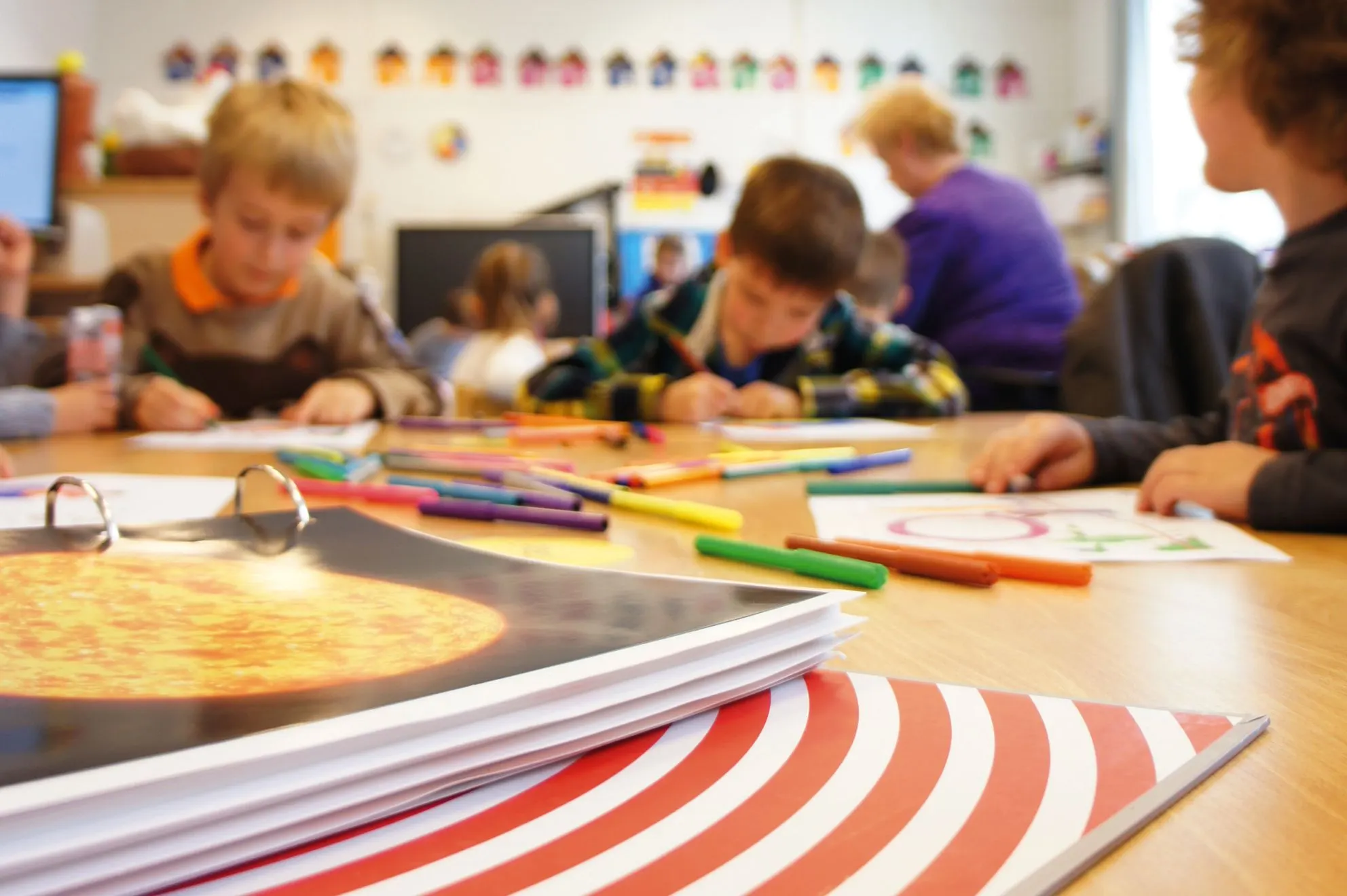 Primary school children drawing at a table with markers, with a ring binder of teaching material in the foreground