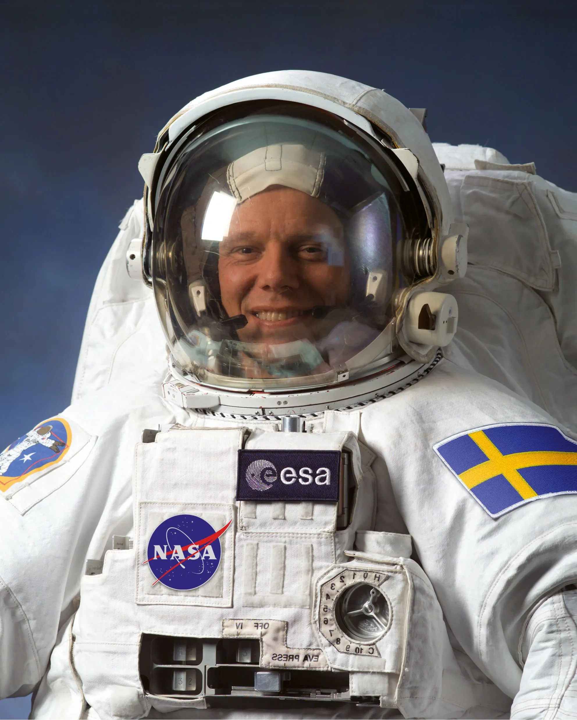 Smiling astronaut in a white spacesuit with NASA, ESA and Swedish flag patches, helmet visor raised
