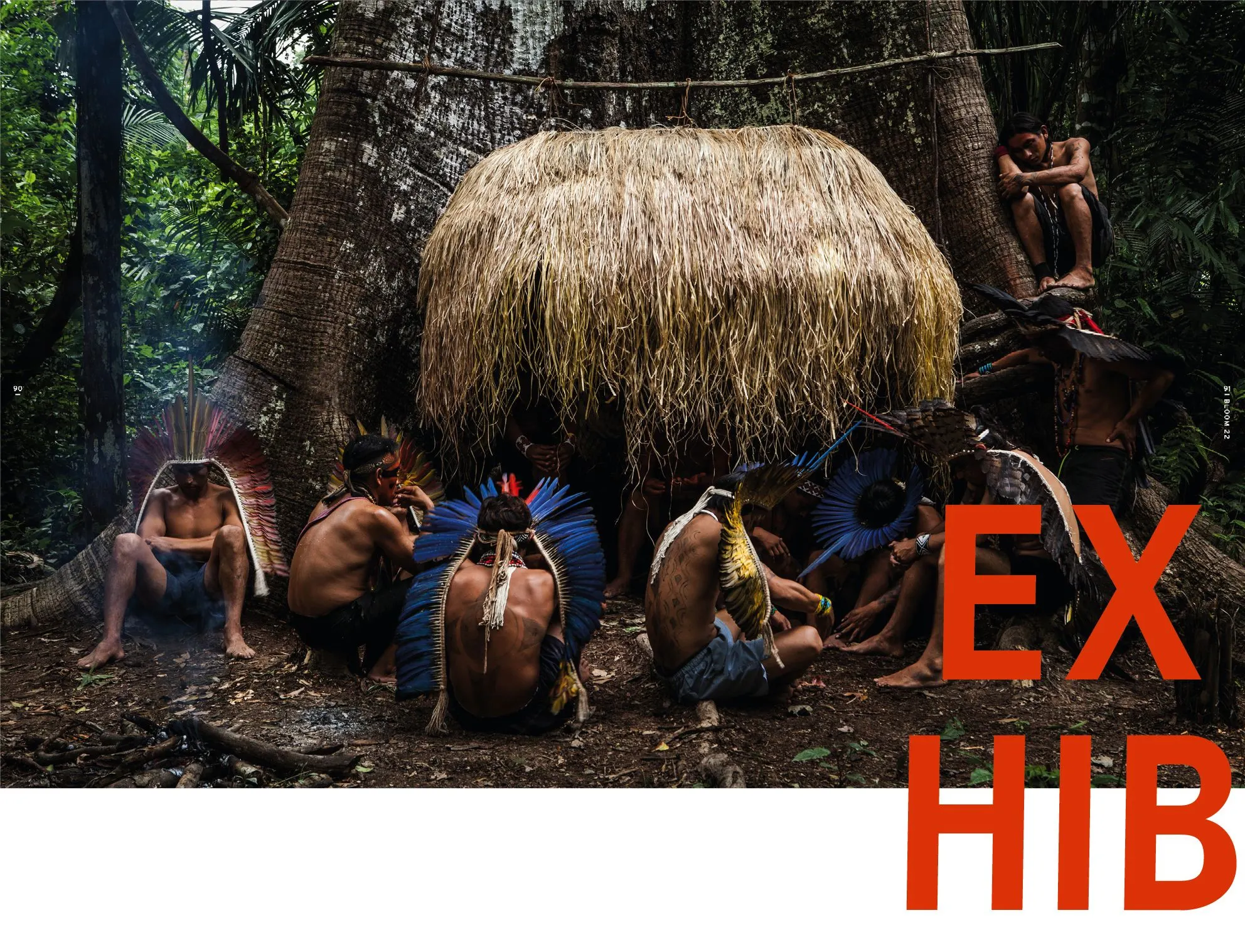 Group of people in feathered headdresses sitting before a large thatched hut in the forest, with red letters EXHIB